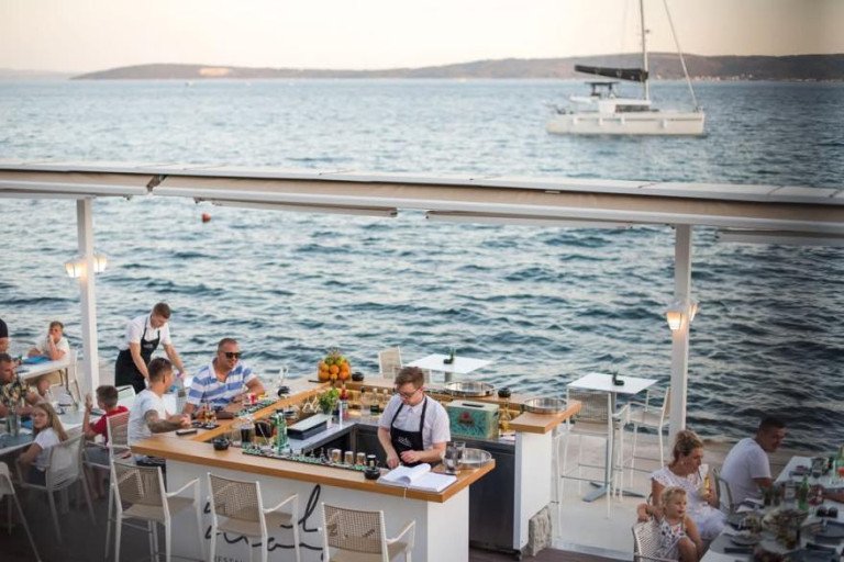 Lively summer atmosphere at the exclusive outdoor terrace and cocktail bar of Mala Mora restaurant right by the sea, with guests, bartenders, and a catamaran in the background