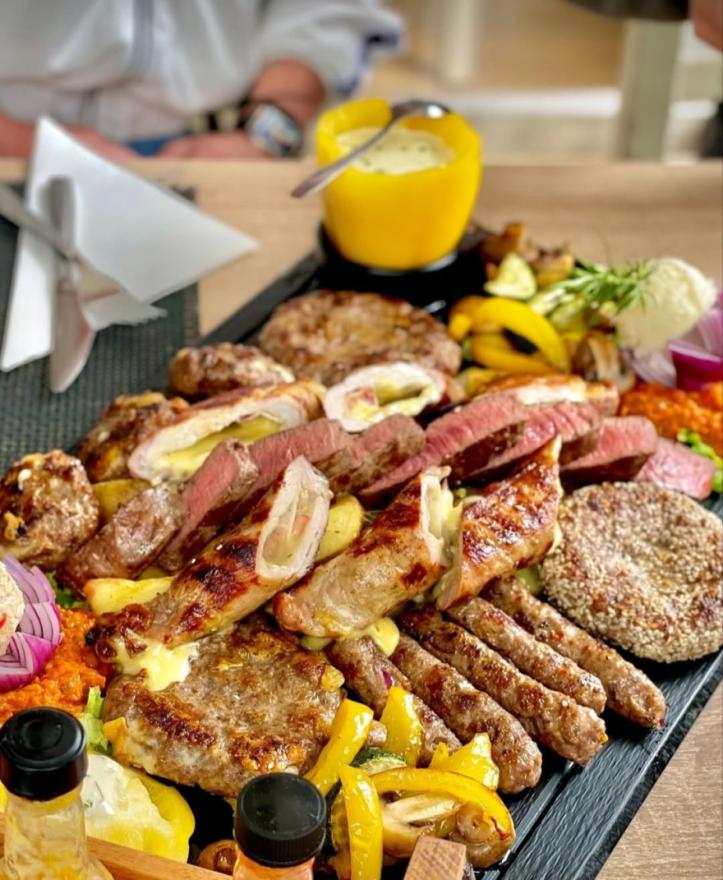 A rich and lavish mixed meat platter featuring various types of grilled meats, ćevapi, steak, stuffed meat cuts, grilled vegetables, and sauces