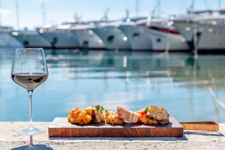 A gourmet appetizer served on a wooden board next to a glass of red wine on the waterfront, with luxury yachts lined up in the background