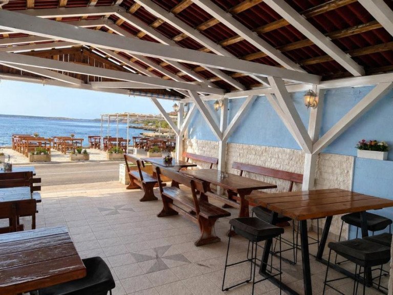Covered terrace of Konoba Amore in Metajna on Pag island with sturdy wooden tables and benches under a white roof structure, offering an open view of the blue sea
