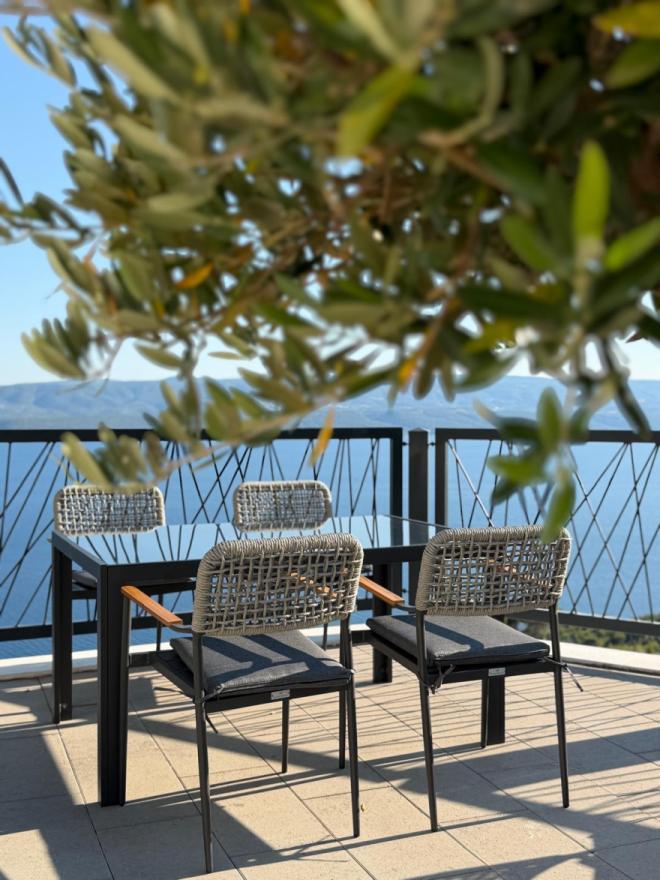A view through olive branches of an elegant outdoor table and woven chairs with dark cushions on the restaurant's terrace overlooking the sea