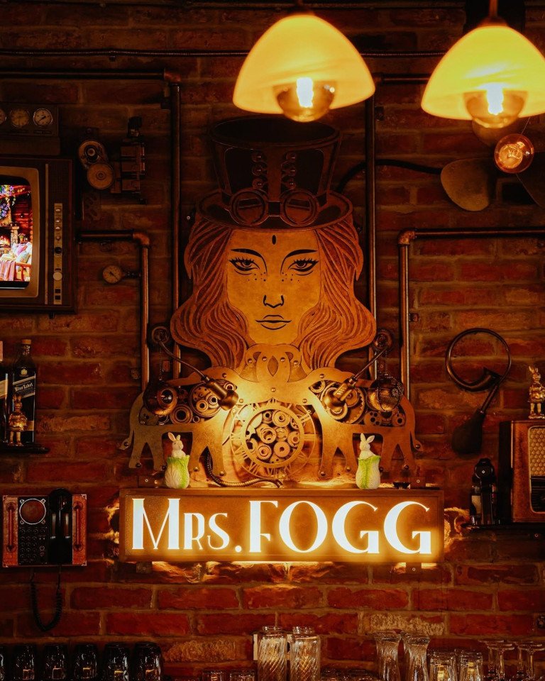 Unikatno uređenje zida u steampunk stilu u baru i pizzeriji Mrs. Fogg s osvijetljenim natpisom, zupčanicima, bakrenim cijevima i ciglenom pozadinom