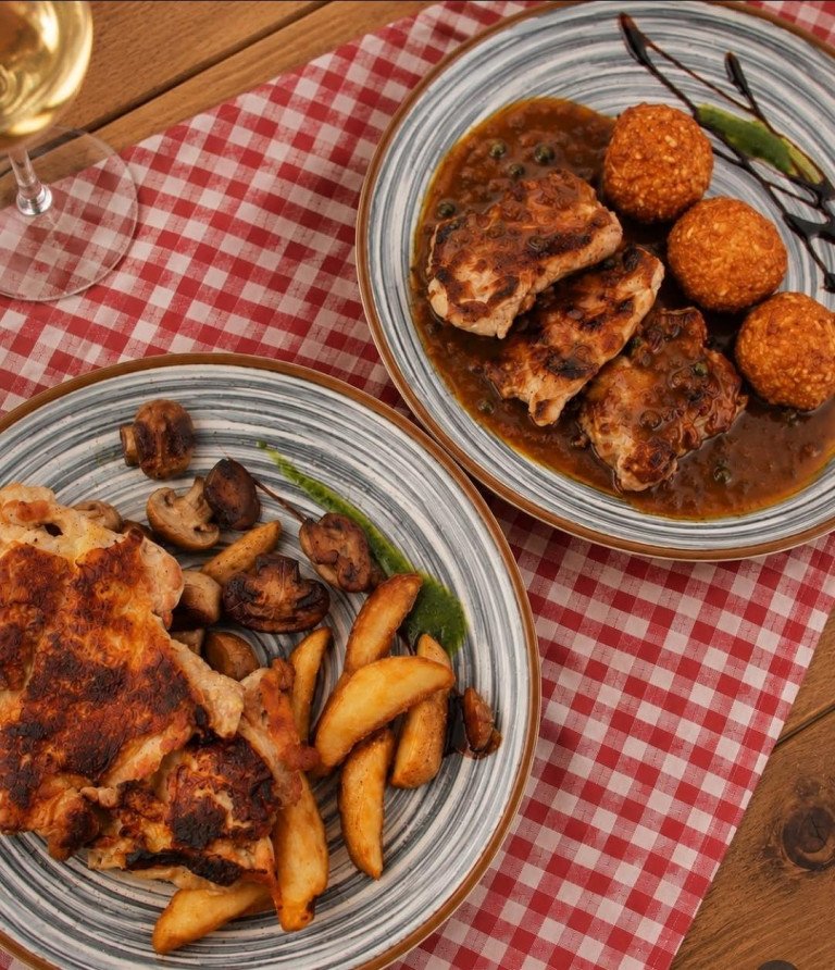 Plated meat dishes including grilled chicken, roasted potatoes, and golden croquettes served on a checkered tablecloth at Konoba Volta in Rijeka