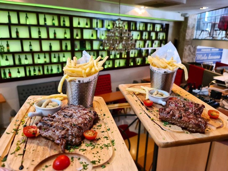 Two grilled steaks served on wooden boards with buckets of french fries, sauces, and cherry tomatoes at Mala Kavana in Split